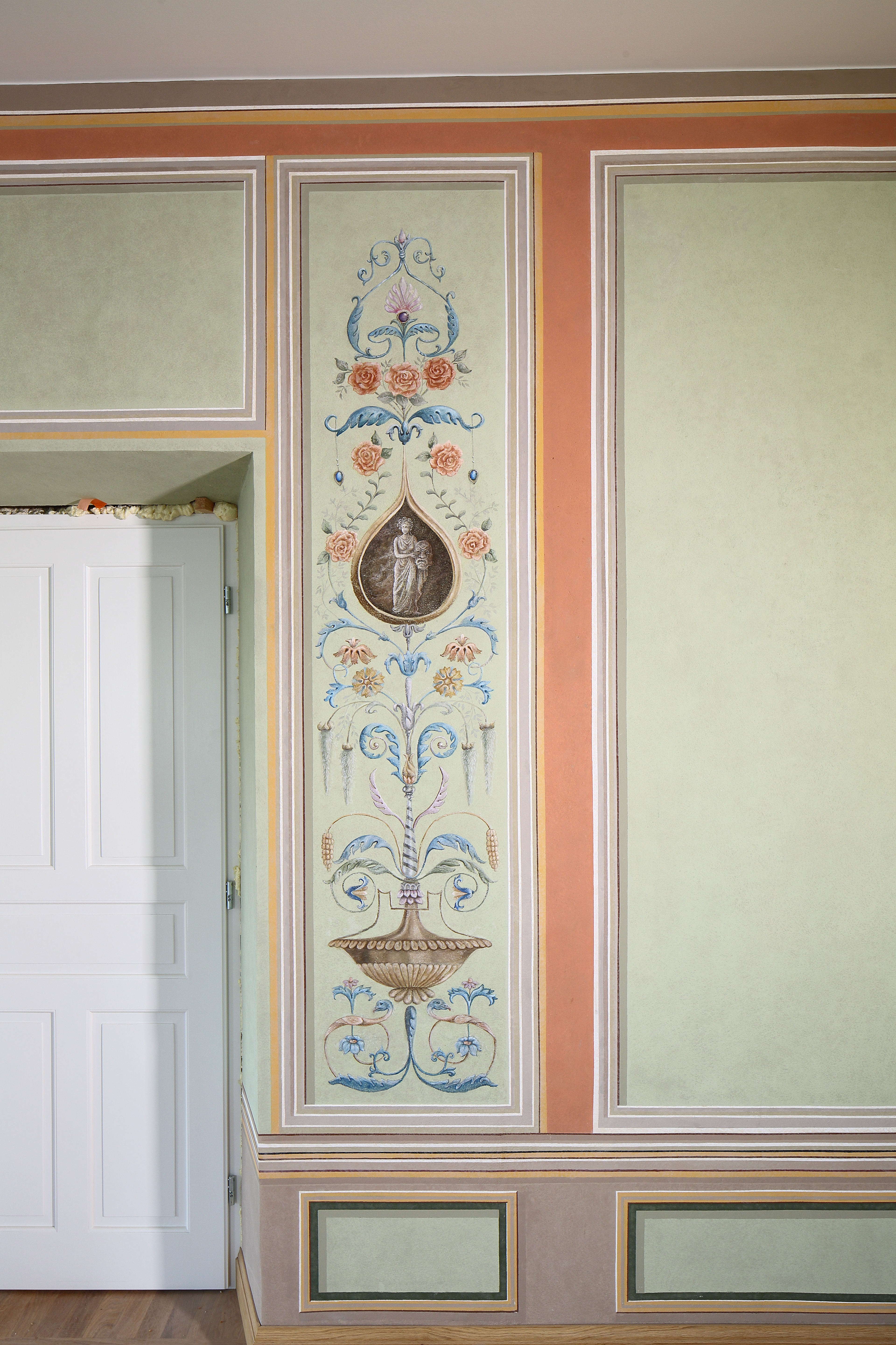 Fragment ściany oraz białych drzwi. Na ścianie namalowane ramki, w jednej z nich wzór kwiatowy. Pozostałe ramki na ścianie są puste.  