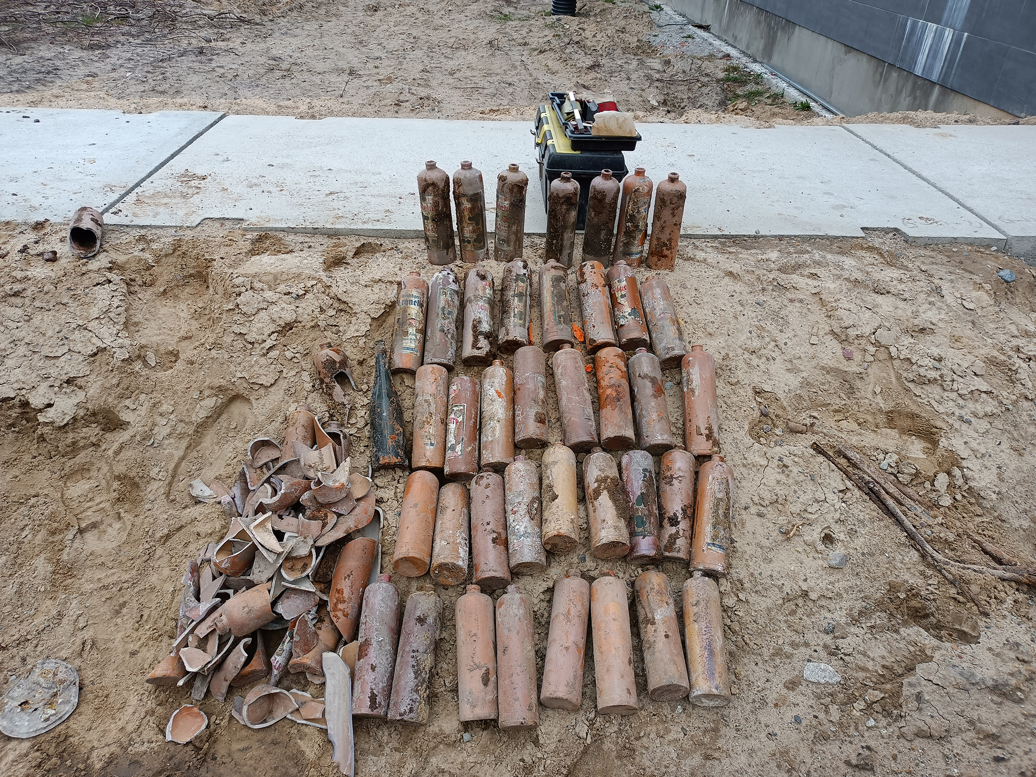 Kilka leżących butelek na piachu, obok  kilka fragmentów potłuczonych butelek. W oddali betonowe płyty ułożone w formie chodnika. 